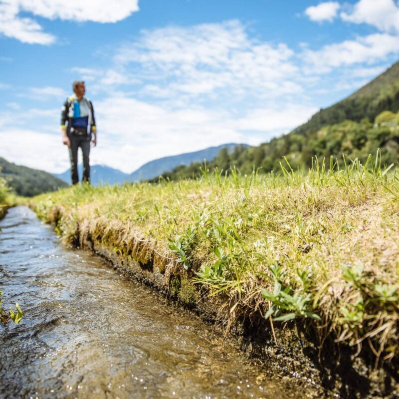 Wanderer neben einem wasserführenden Waal in offener Wiesenlandschaft