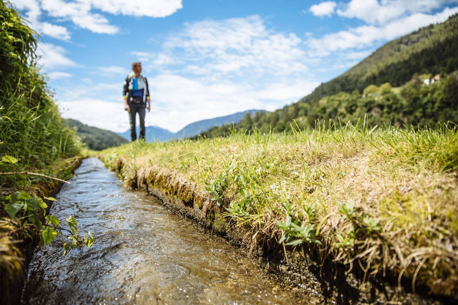 Wanderer neben einem wasserführenden Waal in offener Wiesenlandschaft
